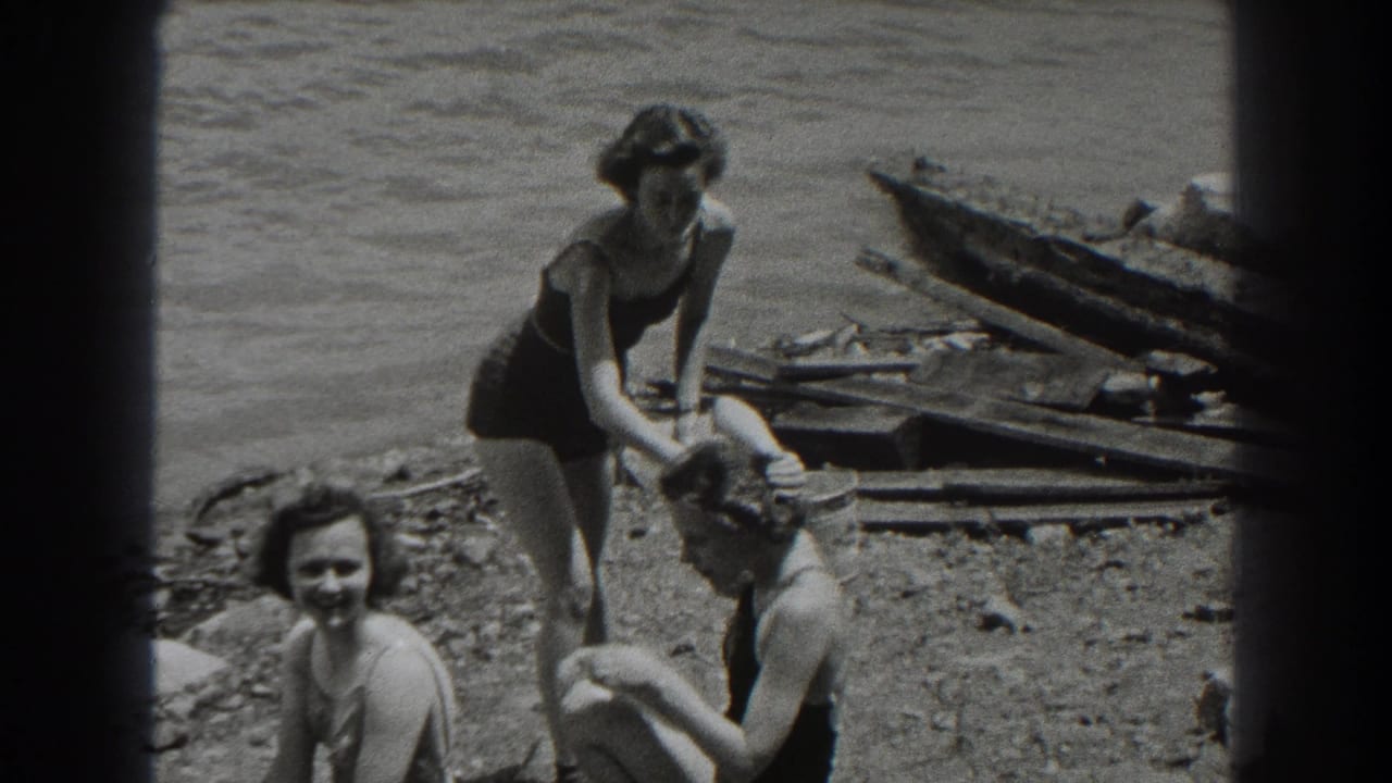1938 New Jersey home-movie frame of three women in swimsuits relaxing by a rocky lake shore, with one adjusting another’s hair.