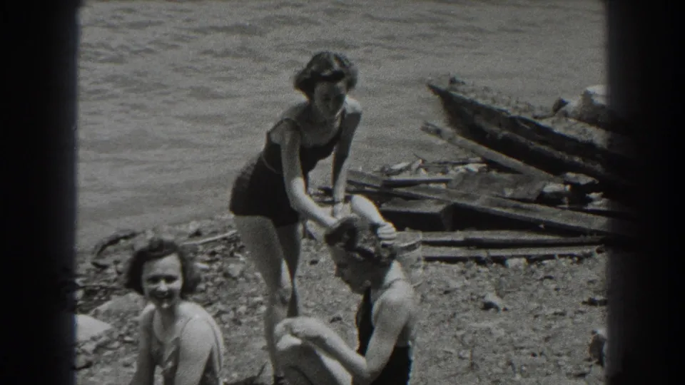 1938 New Jersey home-movie frame of three women in swimsuits relaxing by a rocky lake shore, with one adjusting another’s hair.