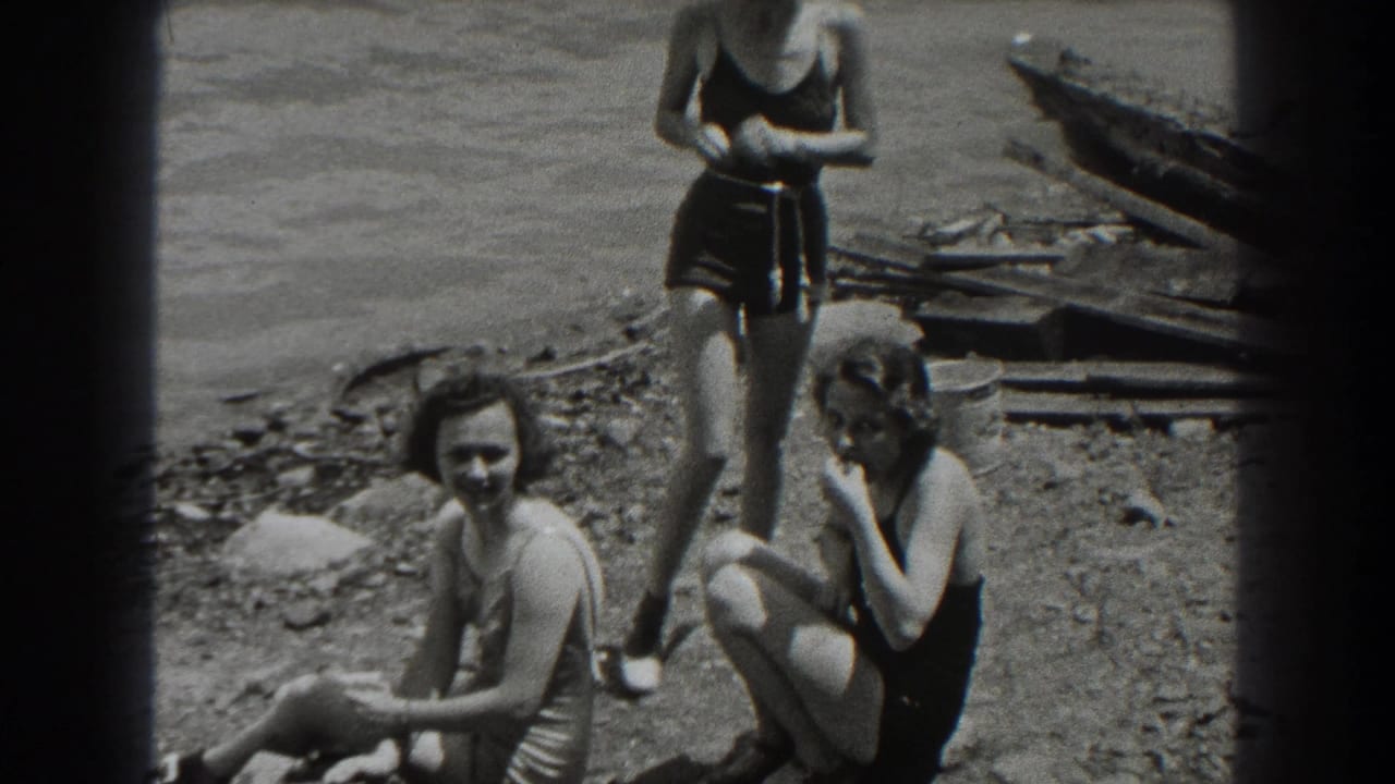 Three women in swimsuits gather on a rocky shore beside lake water and wooden boards.