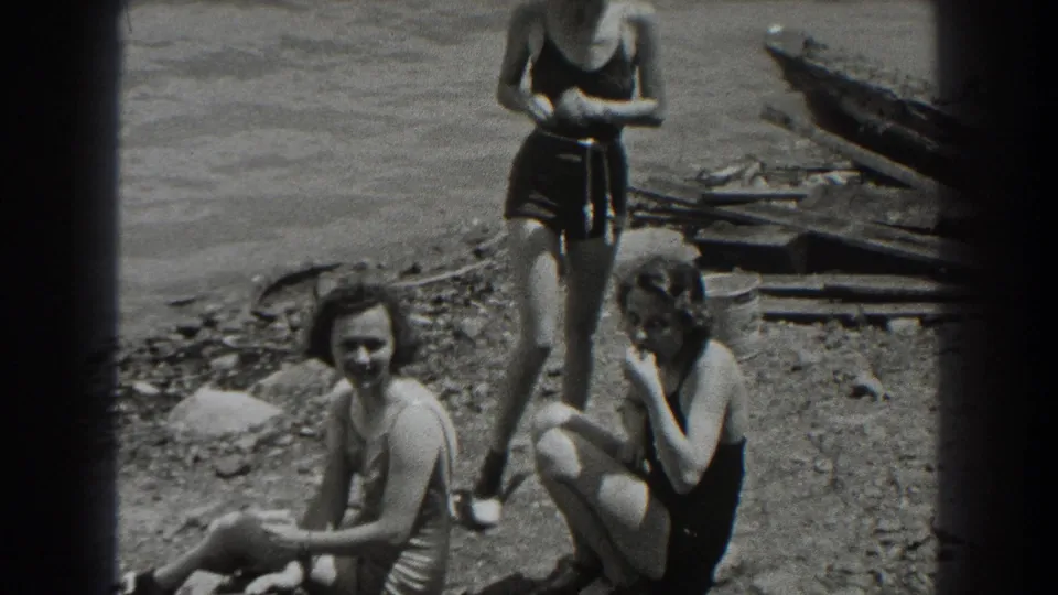 1938 New Jersey lakeside frame showing women in vintage swimsuits, one standing while two sit near water and scattered wooden boards.