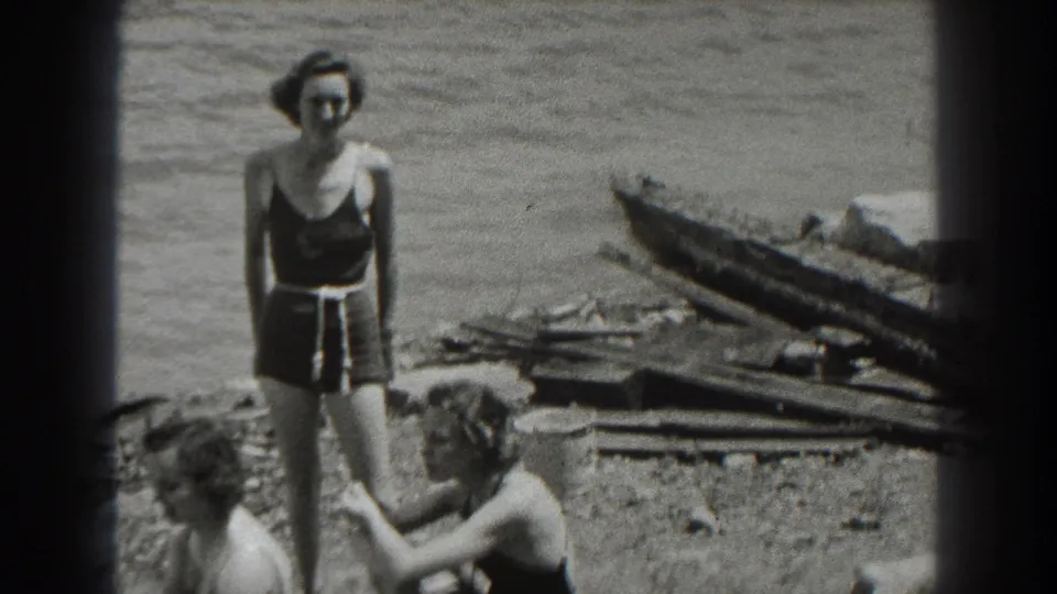 1938 New Jersey lakeshore frame with a woman standing in a dark swimsuit and two seated companions partly visible near boards and water.