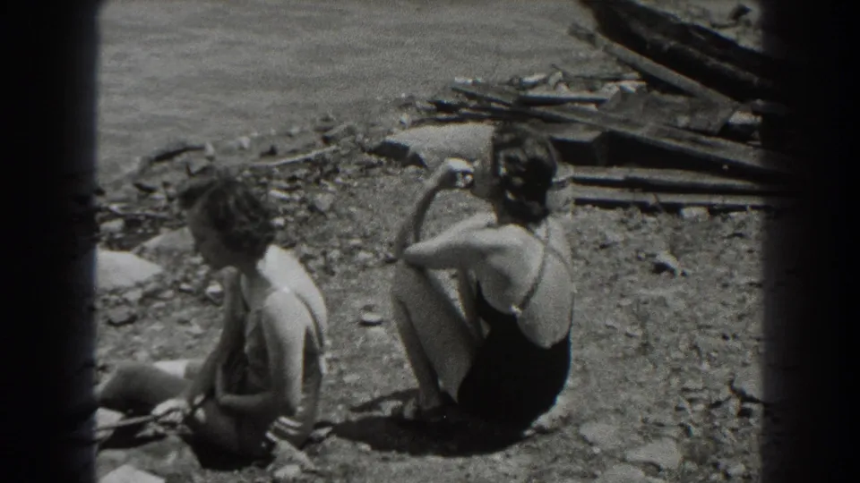 Two adult women in swimsuits sit on a rocky New Jersey shoreline in 1938 home-movie footage, one turned away and one nearer the camera.