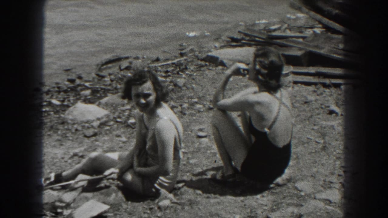 Two women in swimsuits sit on a pebbled beach as one smiles toward the camera.