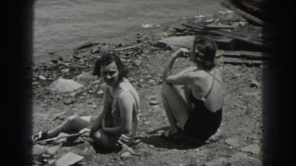 On a rocky New Jersey beach in 1938, one woman smiles toward the camera while another sits with her back turned near scattered driftwood.