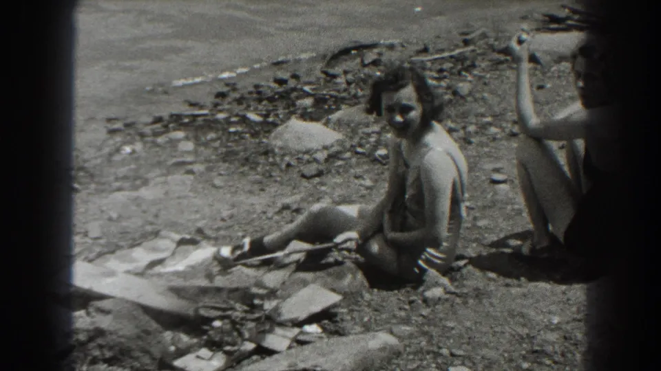 A woman in a swimsuit sits in bright sun on the rocky shore, facing the camera, with another seated figure partly cropped at the right edge.