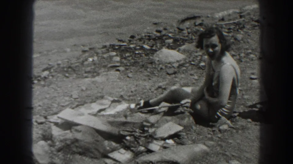 A woman in a swimsuit sits on the pebbled New Jersey shoreline in 1938 footage, with driftwood and shallow water visible behind her.