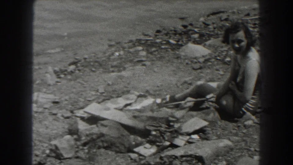 In 1938 New Jersey home-movie footage, a woman in a light swimsuit sits on stones near lake water and debris along the shore.