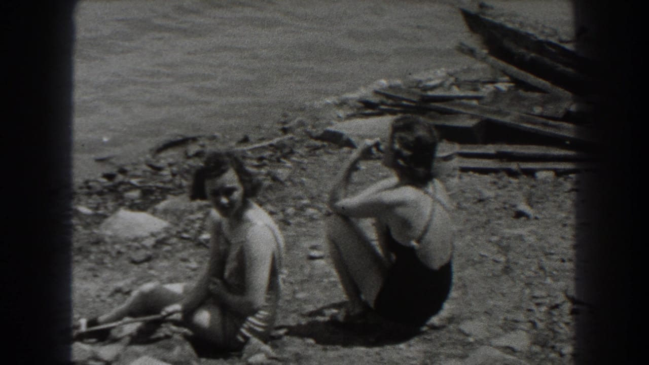 Two women in swimsuits sit on a rocky lakeshore, one holding a cigarette.