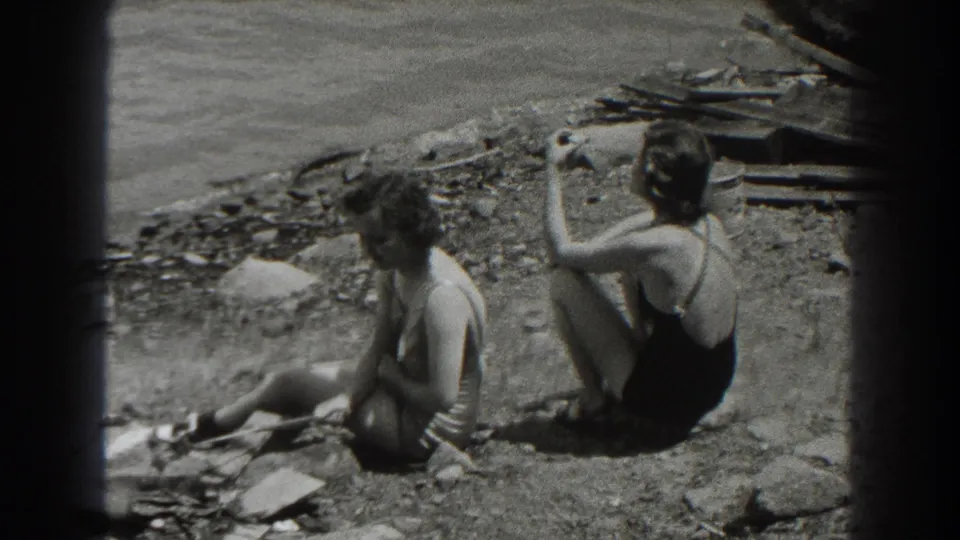 A 1938 New Jersey home-movie frame shows two women seated on a rocky lakeshore, with lake water and scattered boards behind them.
