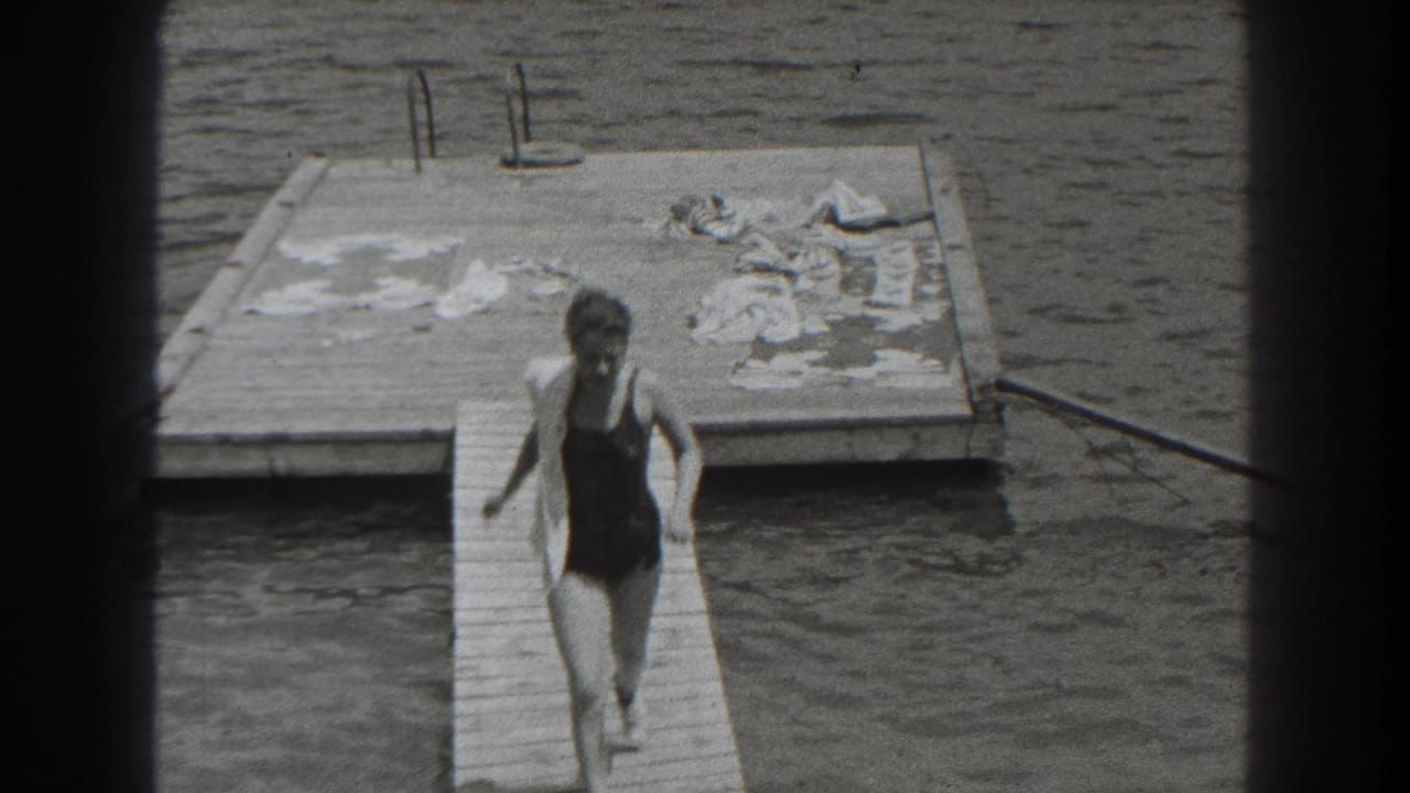 Woman in a one-piece swimsuit runs along a wooden pier toward a floating dock on a lake.