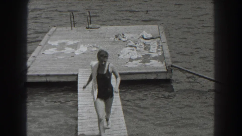 In 1938 New Jersey home-movie footage, a woman in a dark one-piece swimsuit runs down a wooden pier beside calm lake water.
