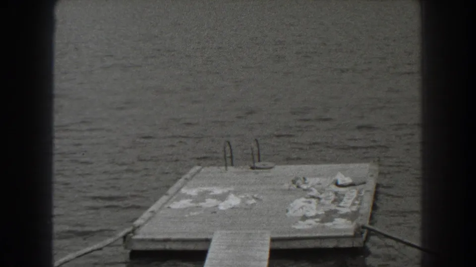 1938 New Jersey home-movie frame with a dock platform, ladder rails, rippled lake water, and wooded shore beyond.