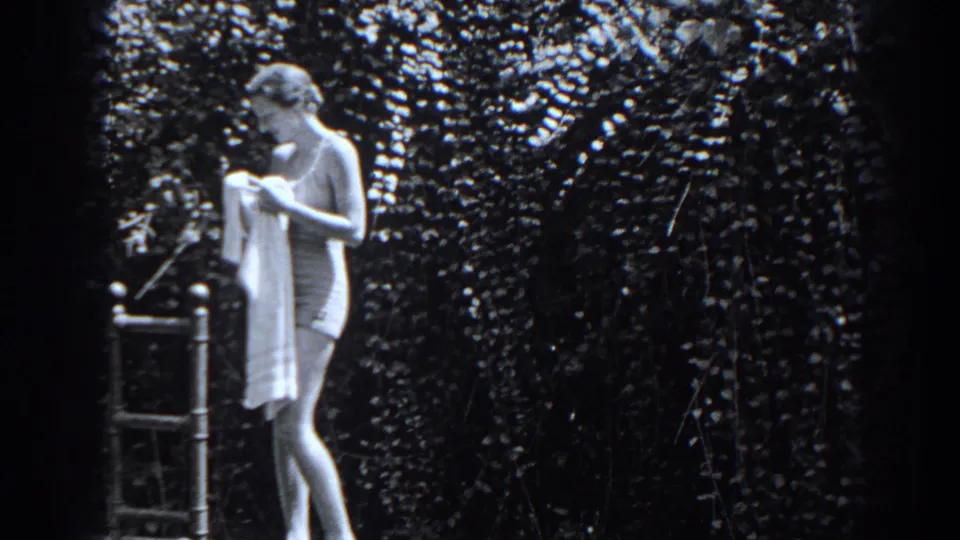 In 1938 New Jersey home-movie footage, a young woman in a bathing suit holds a towel beside a metal ladder and dense foliage.