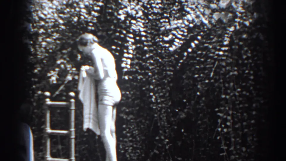 In supported 1938 New Jersey footage, the woman bends over a towel near a metal ladder, with dense ivy-like foliage behind her.