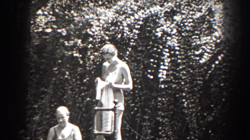 1938 New Jersey frame of a standing woman with a towel near a ladder and a seated woman before a leafy hedge.