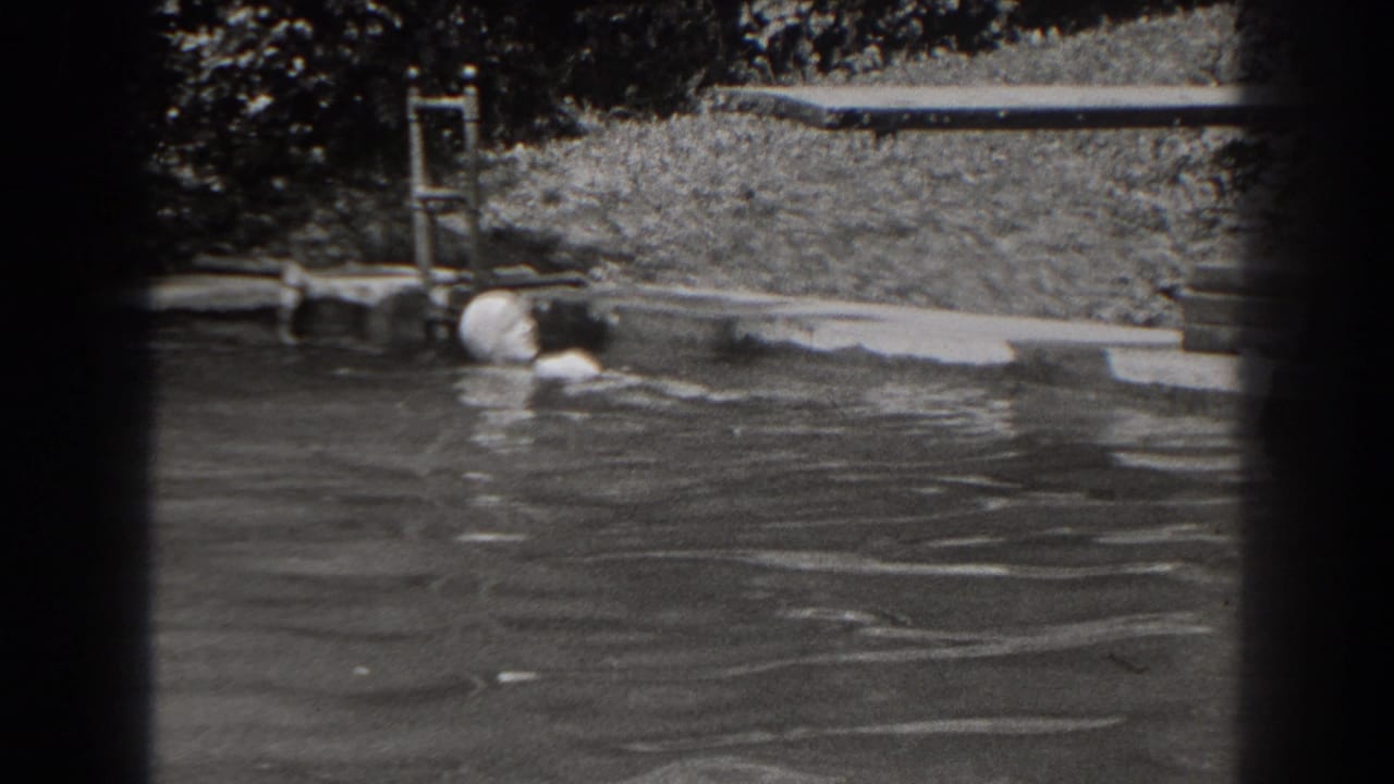 1938 New Jersey frame showing a woman swimming across a backyard pool with the ladder, lawn, and dense plants in view.