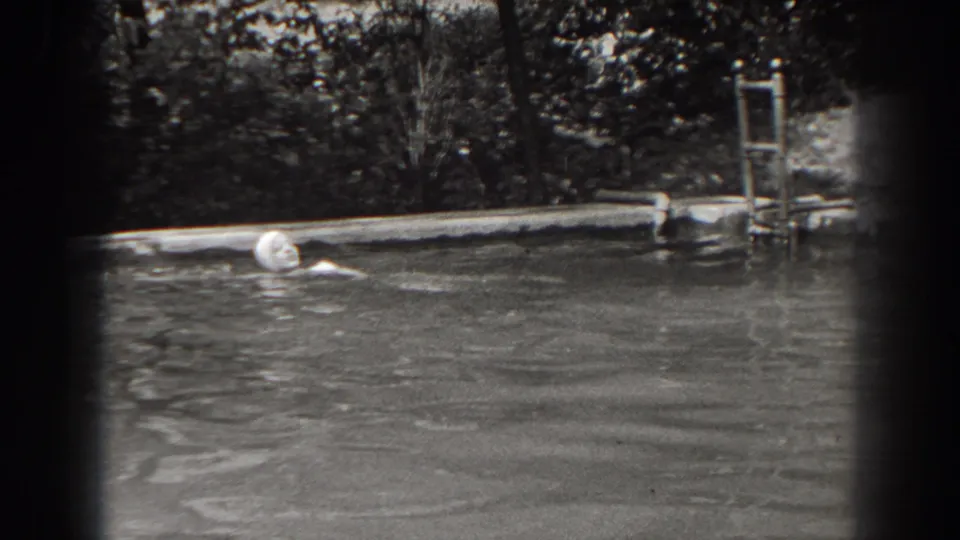 1938 New Jersey 8mm frame showing a woman in a light swim cap swimming across a backyard pool with a ladder and leafy background.