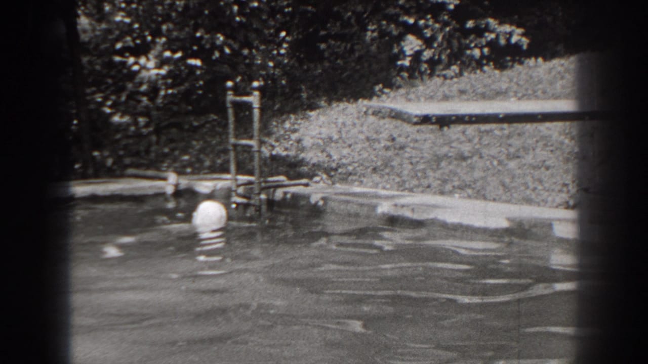 1938 New Jersey home-movie frame with the swimmer's capped head near the pool edge, metal ladder, grass, and trees visible behind.