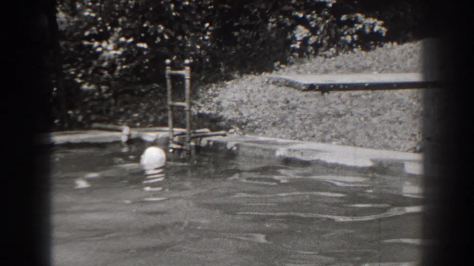 1938 New Jersey home-movie frame with the swimmer's capped head near the pool edge, metal ladder, grass, and trees visible behind.