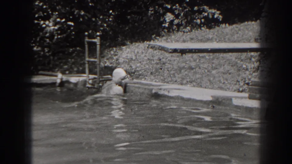 1938 New Jersey home-movie frame of a woman upright in the pool near the coping, with a ladder, grass, and trees in the background.