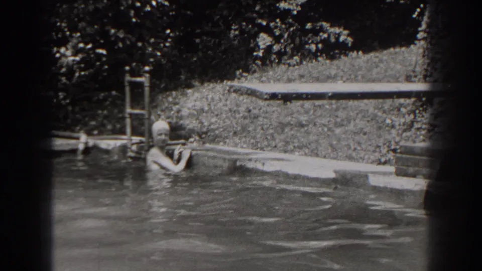 In 1938 New Jersey home-movie footage, a woman in a swim cap pauses at the edge of an outdoor pool near a ladder and diving board.