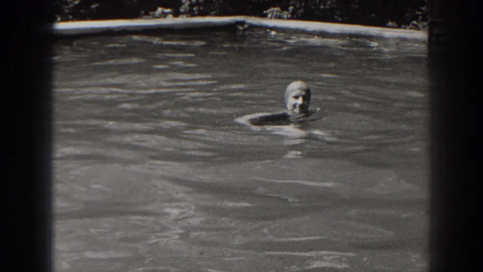 A woman in a swim cap faces the camera while floating in gray pool water, consistent with the 1938 New Jersey home-movie clip.