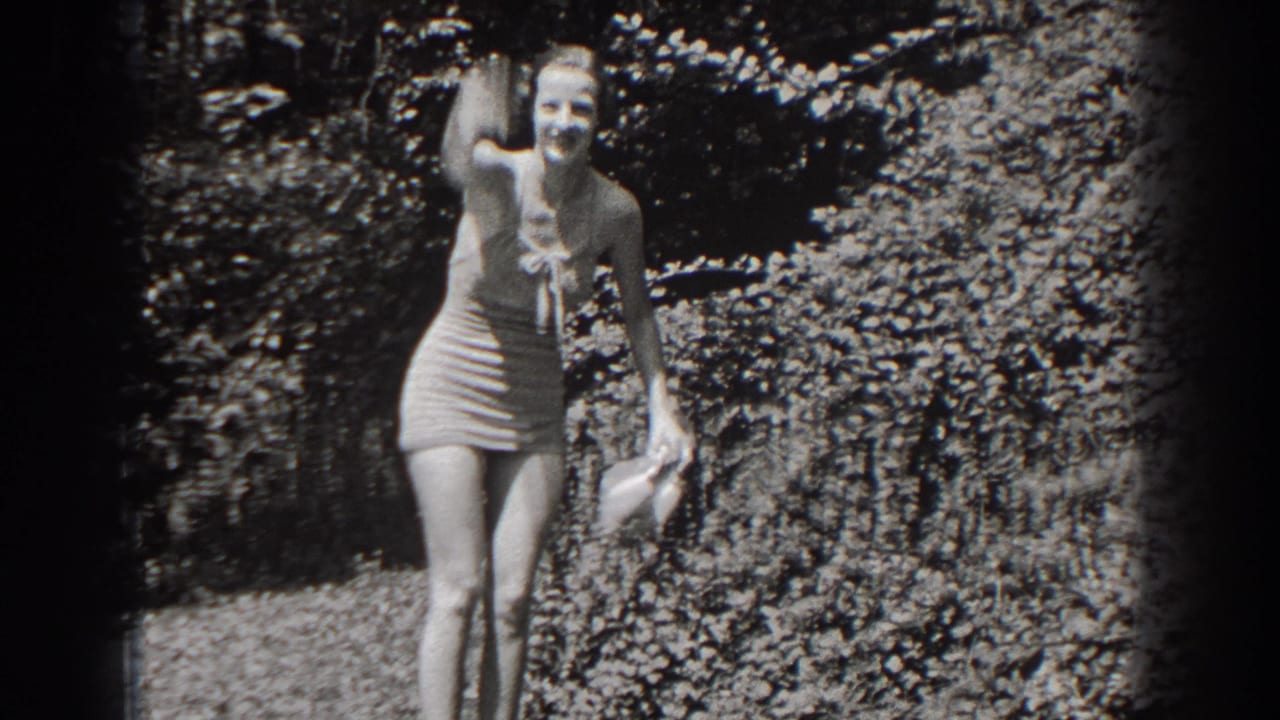 Woman in a light one-piece swimsuit waves while standing in a leafy garden.