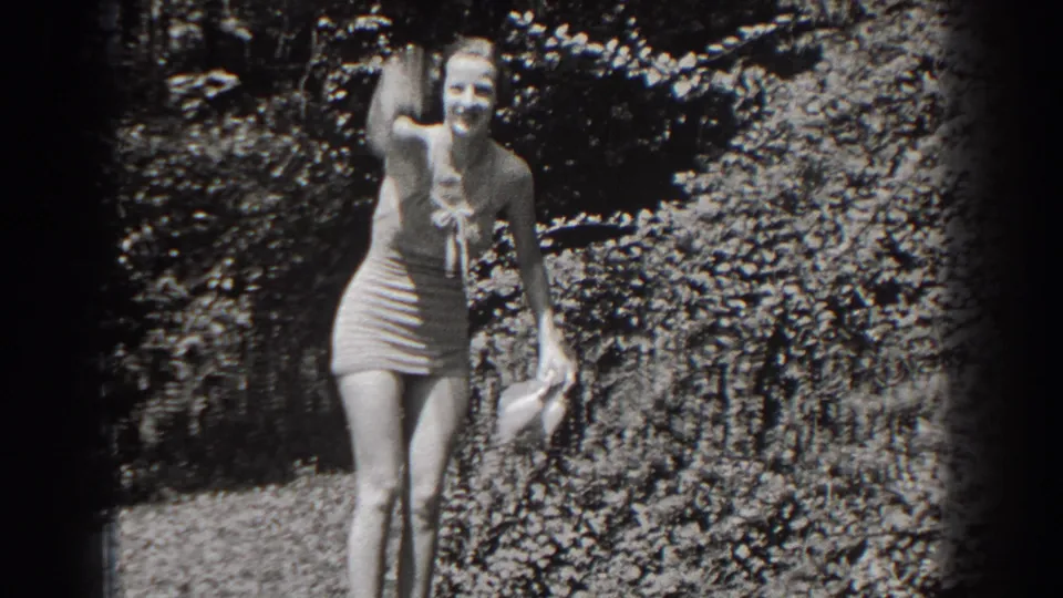 In 1938 New Jersey home-movie footage, a woman in light swimwear stands in sunlit foliage and waves toward the camera.