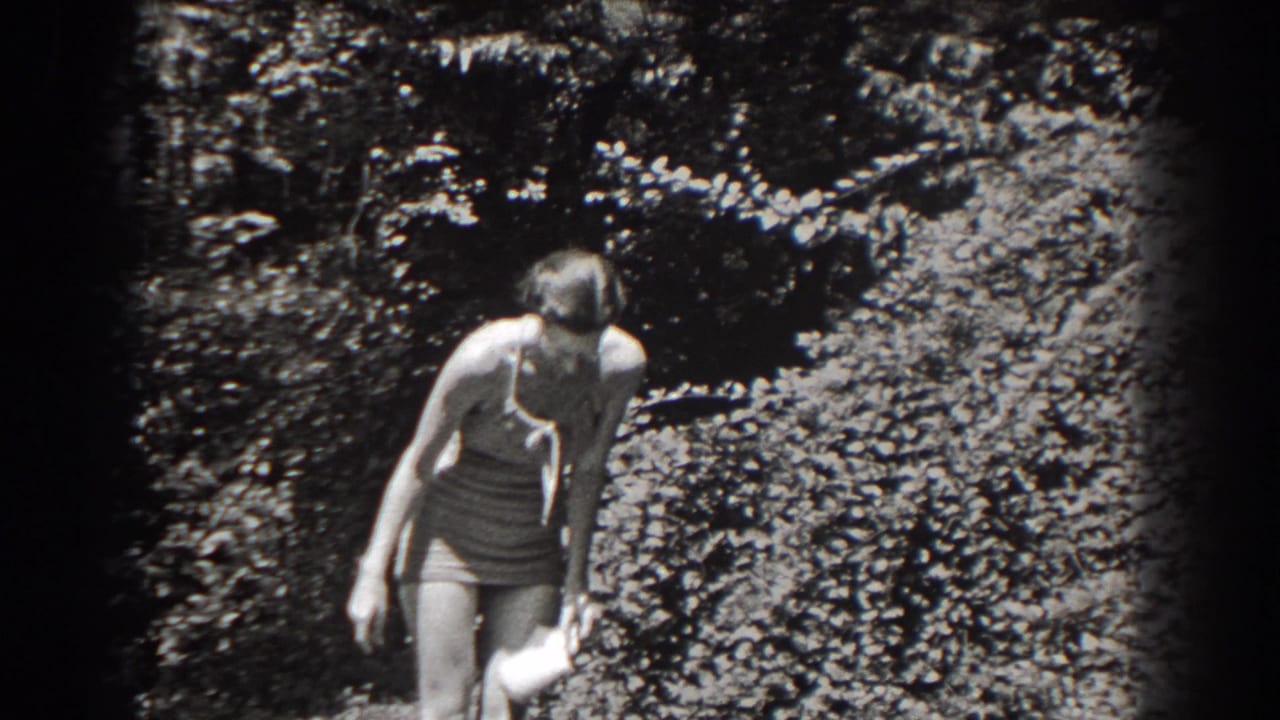 Woman in swimwear looks down while holding a small light object near garden shrubs.
