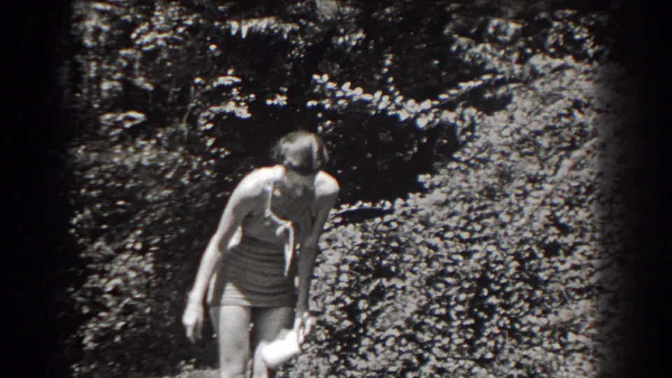 The woman in light swimwear looks downward in a sunlit New Jersey garden, holding a small pale object at her side.