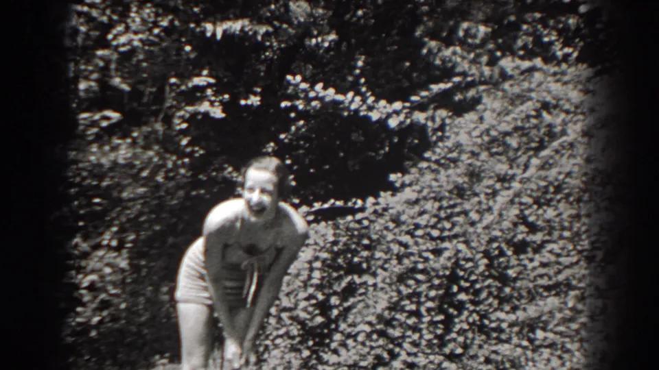 A woman in 1938 New Jersey home-movie footage bends forward smiling, framed by sunlit garden foliage.