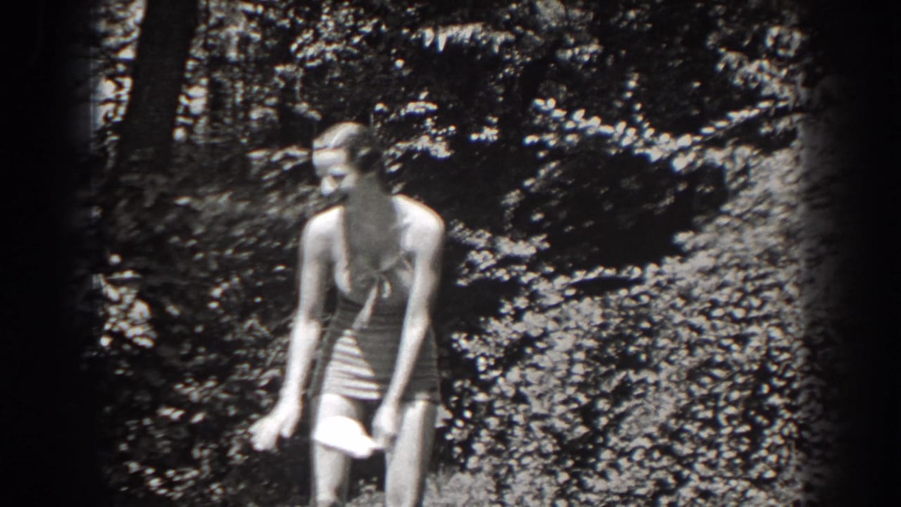 Woman in a light striped swimsuit stands in front of leafy brush holding a hat.