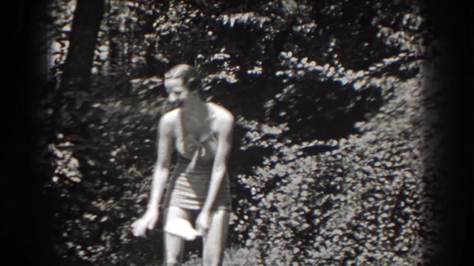 In metadata-supported 1938 New Jersey footage, a woman in light striped swimwear stands before dense foliage holding a hat.