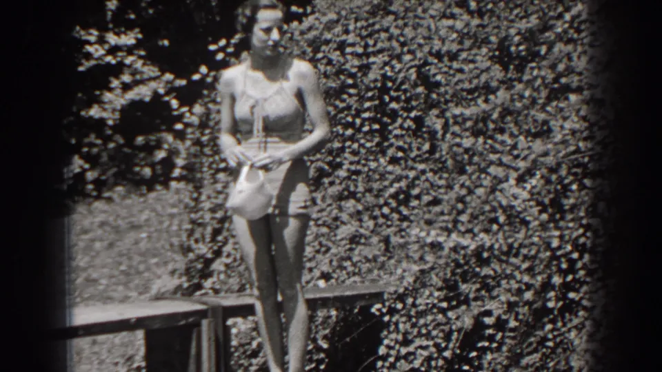 In the clearest frame, the woman stands near dense New Jersey foliage and a low rail, holding her hat in front of her swimsuit.