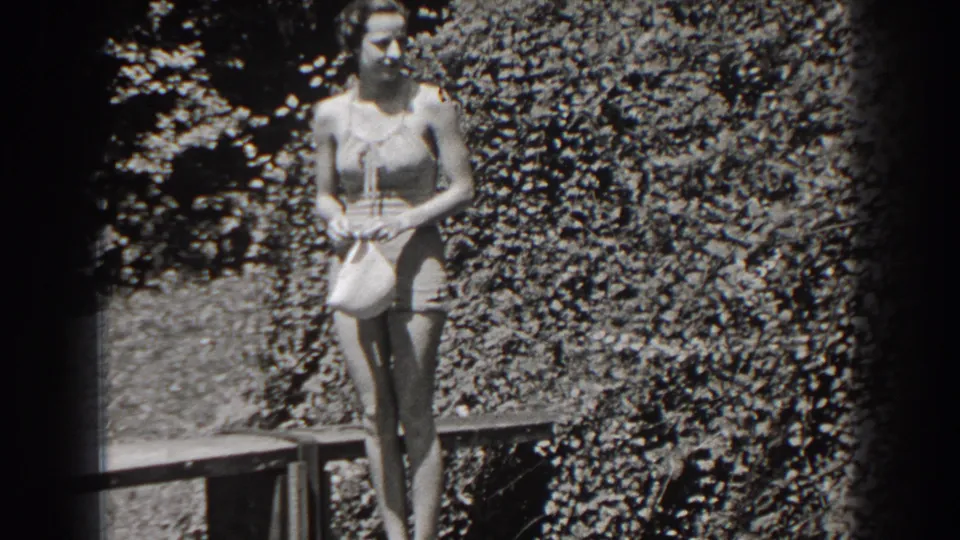 In 1938 New Jersey home-movie footage, a woman in a one-piece bathing suit stands by foliage holding a swim cap.