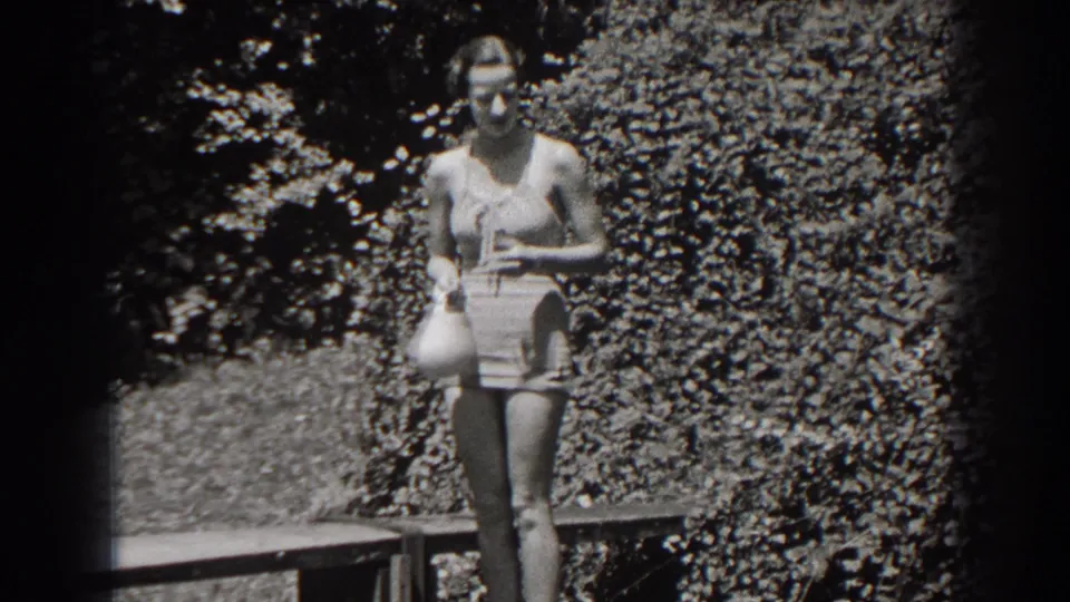 The woman remains posed in the New Jersey outdoor scene, holding a swim cap against a leafy background.