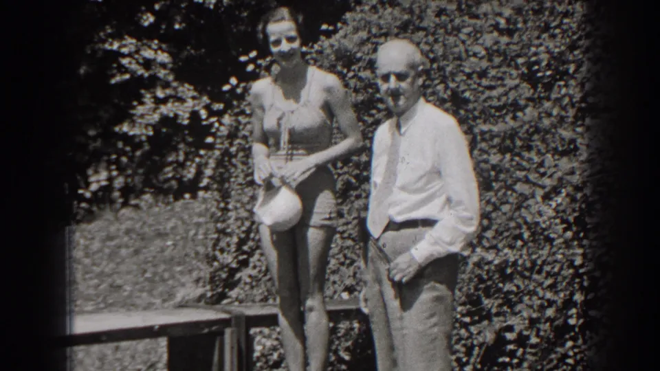 An older man joins the woman in the 1938 New Jersey home-movie frame, both standing near the wooden deck and foliage.