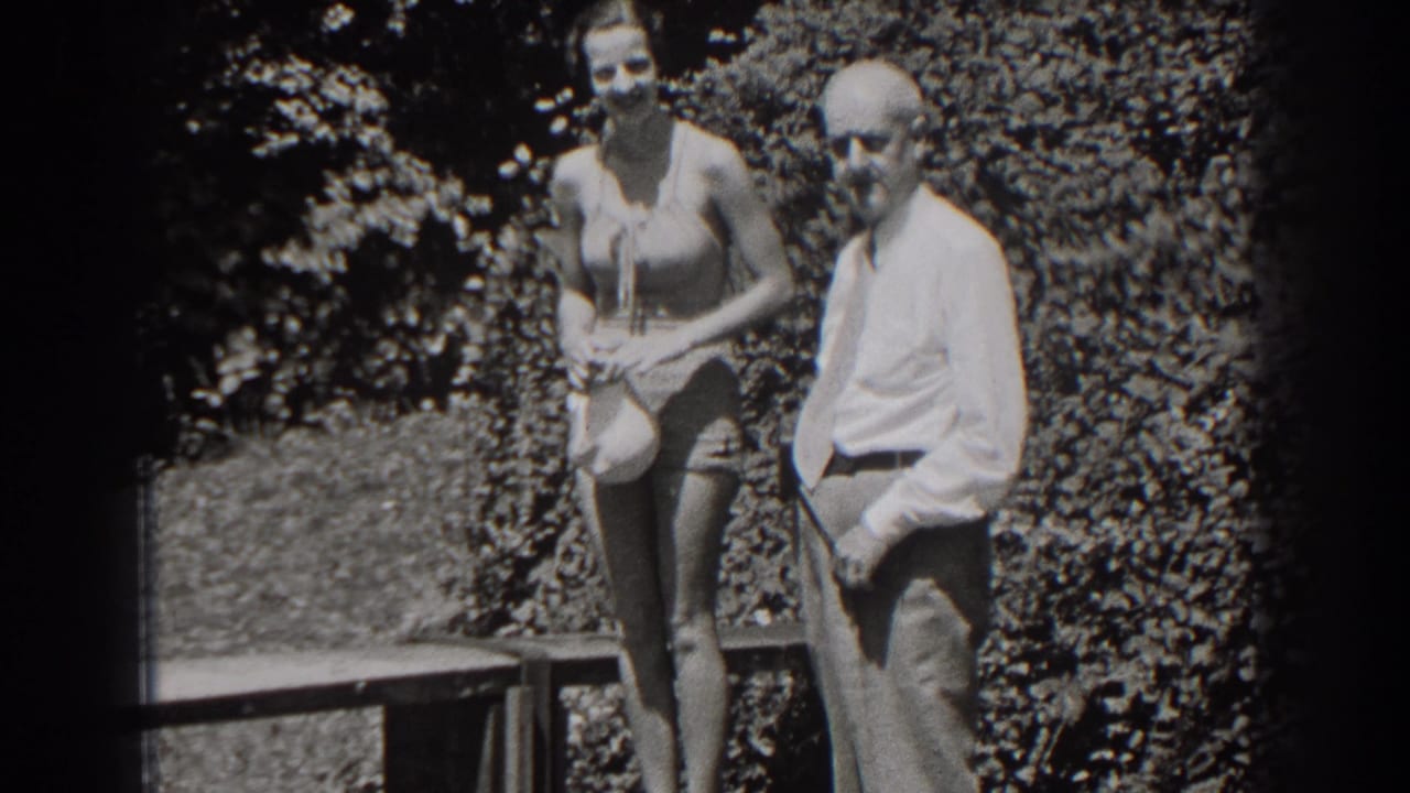 Woman in a swimsuit holds a light cap beside a man in a dress shirt in a garden.