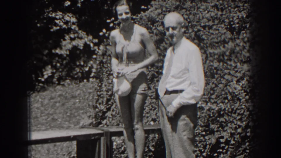 In 1938 New Jersey home-movie footage, a woman in a one-piece swimsuit stands near a man in a dress shirt and tie beside a wooden bench.