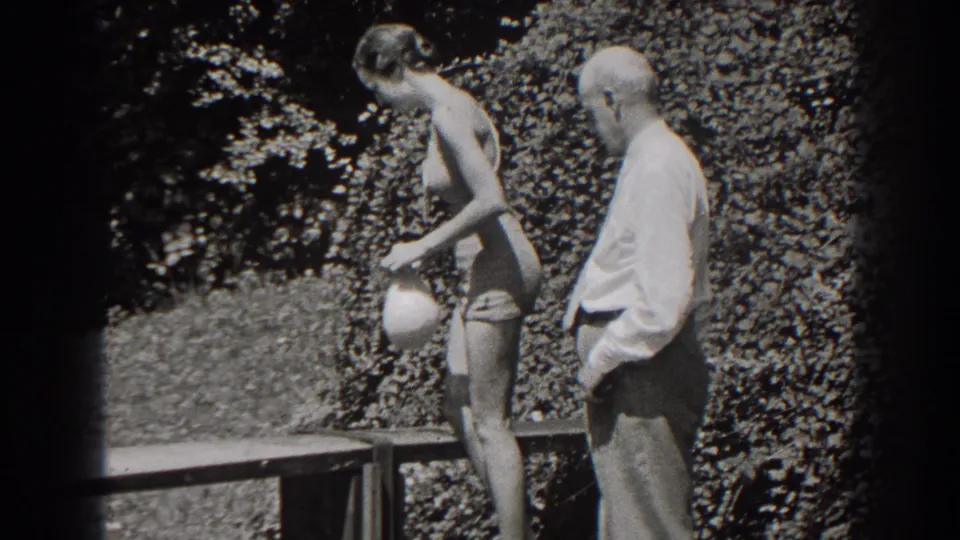 The woman turns toward a wooden bench with a cap in hand while the man in shirt and tie stands beside her in the garden.