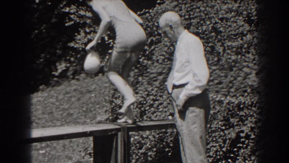 The woman in the one-piece swimsuit steps up onto the wooden bench as the man in a dress shirt and tie remains close by in the garden.