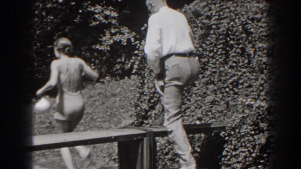 The woman moves away from the bench with her back to camera while the man follows, both framed by grass and dense shrubs.