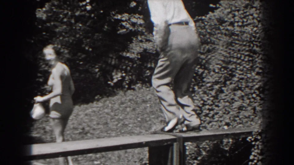 In 1938 New Jersey home-movie footage, a woman in a swimsuit holds a white cap as an older man stands partly cropped on a wooden platform.