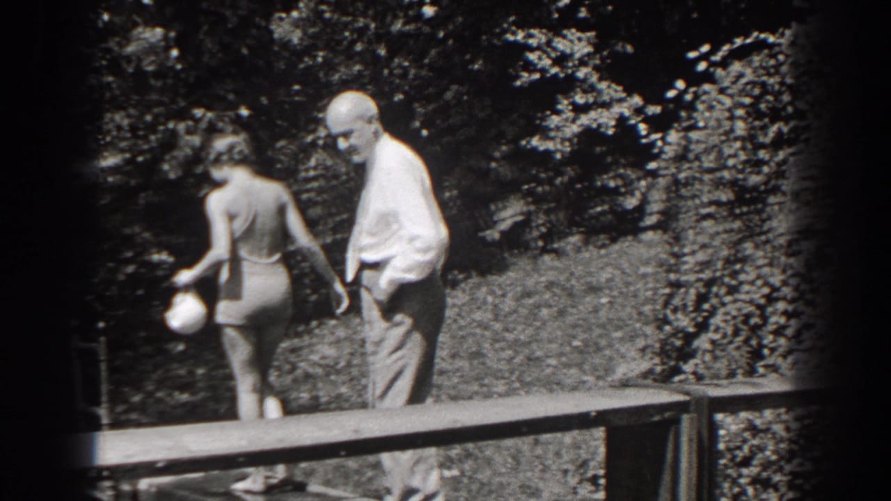 In 1938 New Jersey, an older man in a light shirt and trousers stands near a woman in a swimsuit by a wooden platform and trees.
