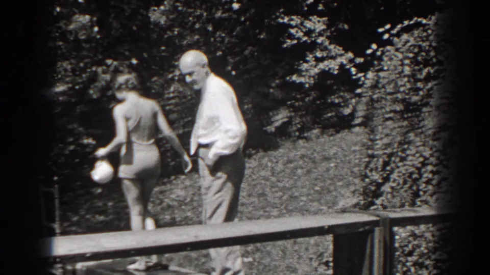 In 1938 New Jersey, an older man in a light shirt and trousers stands near a woman in a swimsuit by a wooden platform and trees.