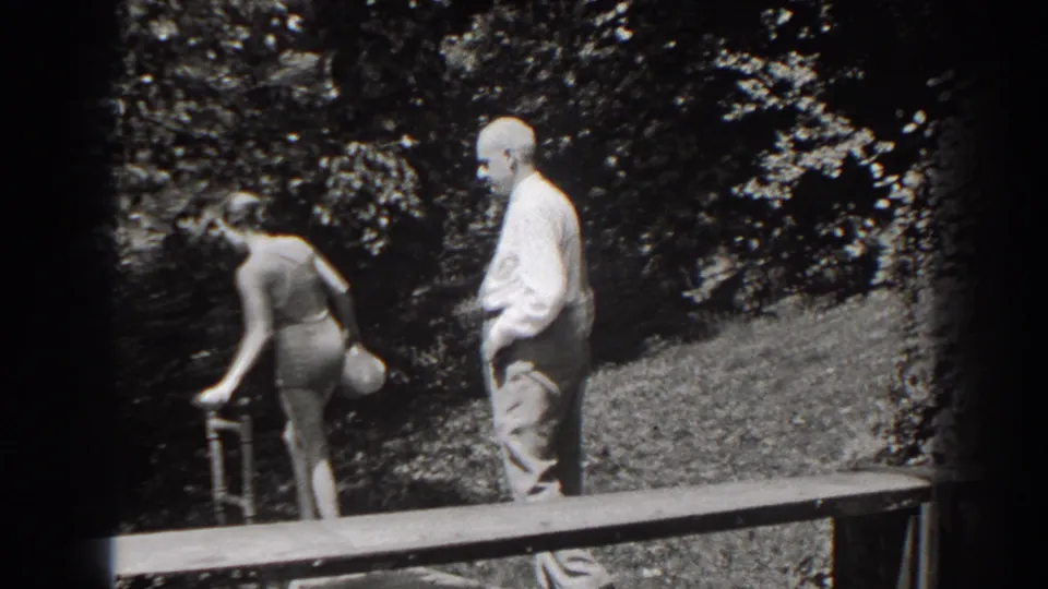 The woman bends toward a small fixture near the platform as the older man watches from the sunlit wooden edge.