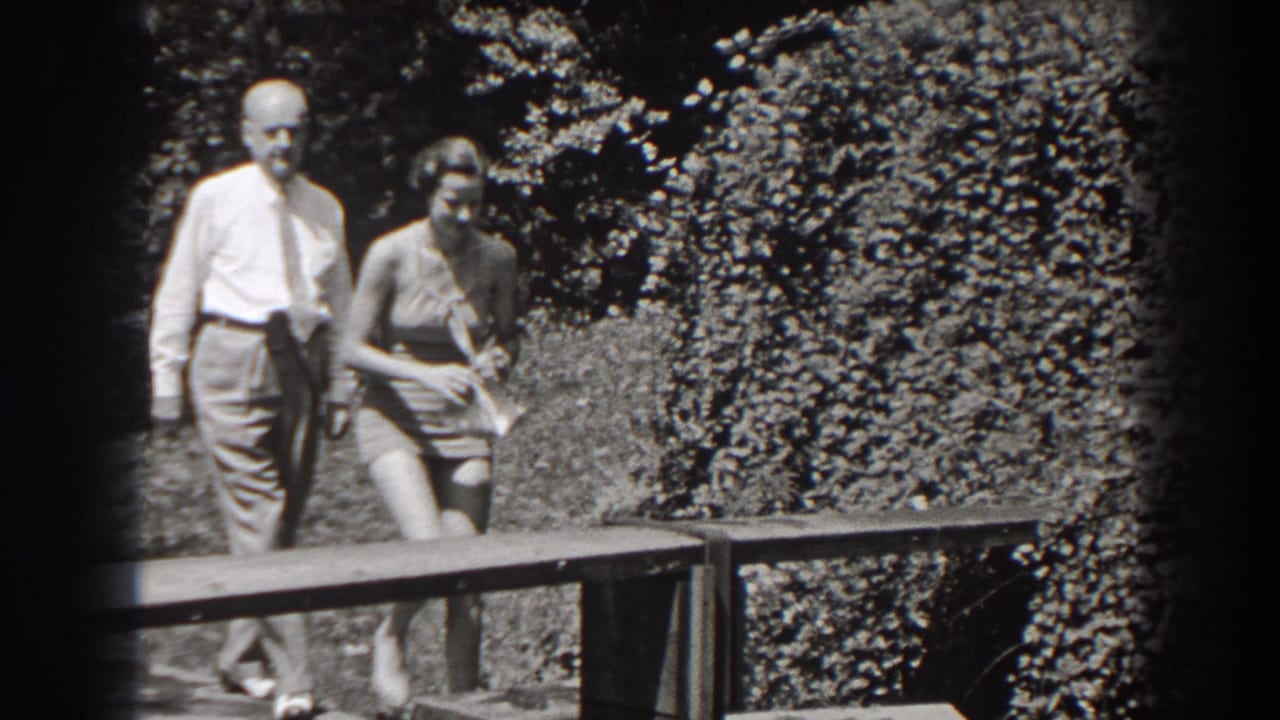 Older man and young woman walk beside a wooden bench in a leafy garden.
