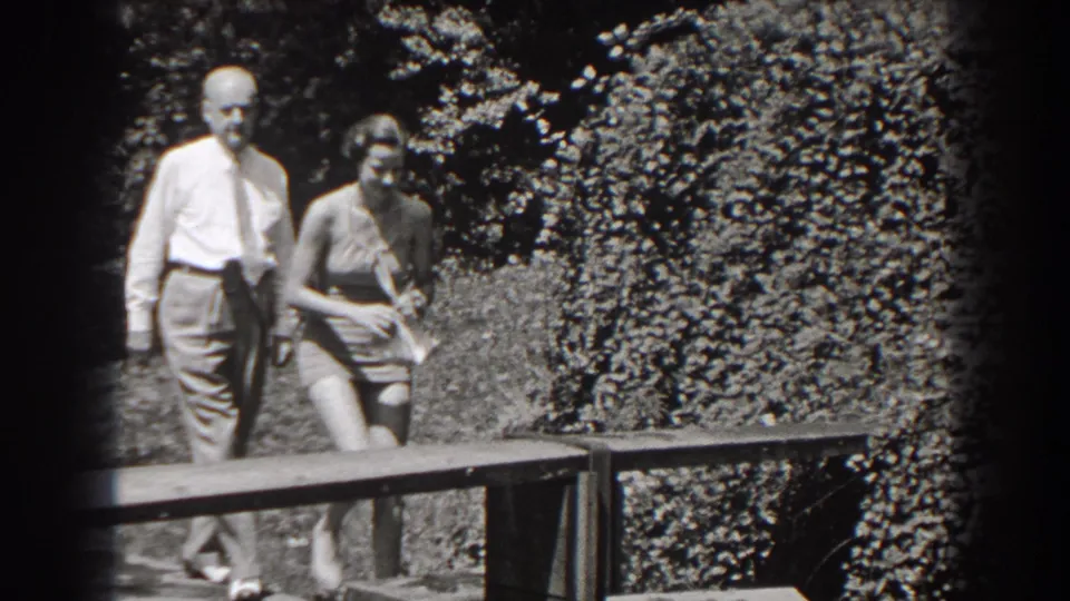 In 1938 New Jersey home-movie footage, an older man in a shirt and tie walks with a young woman in a swimsuit near a garden bench.