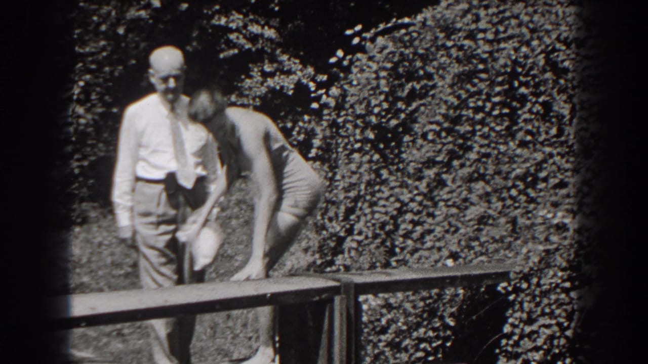 The young woman bends toward a bench or object while the older man stands close by in a leafy New Jersey garden setting.