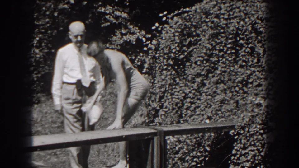 The young woman bends toward a bench or object while the older man stands close by in a leafy New Jersey garden setting.